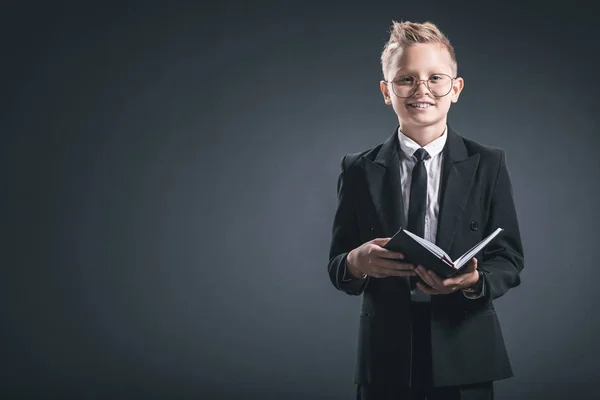 Smiling boy in businessman suit and eyeglasses with notebook on grey backdrop — Stock Photo