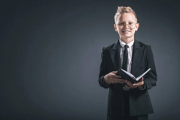 Menino sorridente em terno de empresário e óculos com caderno em pano de fundo cinza — Fotografia de Stock