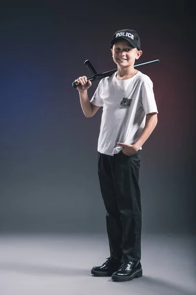 Niño sonriente en camiseta blanca y gorra uniforme oficial de policía con porra en el fondo oscuro - foto de stock