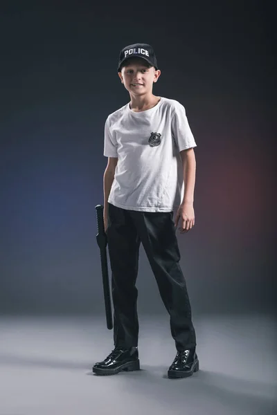 Niño sonriente en camiseta blanca y gorra uniforme oficial de policía con porra en el fondo oscuro - foto de stock