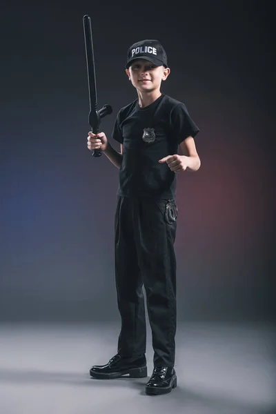 Preteen boy in policeman uniform with truncheon on dark backdrop — Stock Photo