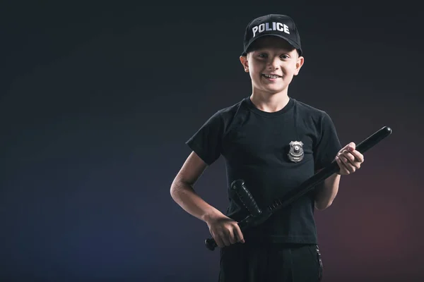 Niño preadolescente sonriente en uniforme de policía con porra en el fondo oscuro - foto de stock
