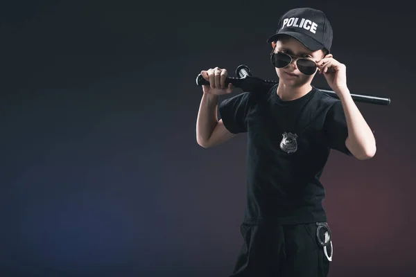 Portrait of boy in policeman uniform and sunglasses with truncheon on dark background — Stock Photo