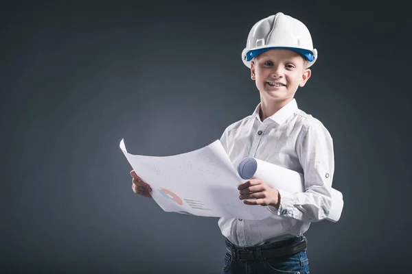 Retrato de niño sonriente en casco de arquitecto sosteniendo planos sobre fondo oscuro - foto de stock