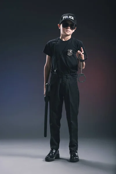 Boy in policeman uniform and sunglasses with handcuffs on dark background — Stock Photo