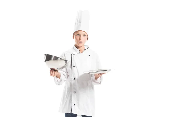 Chocado chico en uniforme de chef y sombrero sosteniendo bandeja de servir vacía aislado en blanco - foto de stock