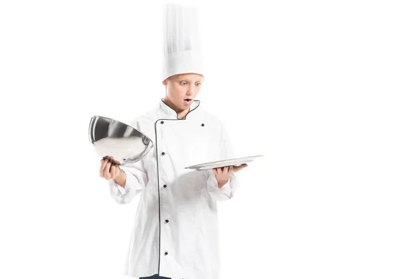 Chocado chico en uniforme de chef y sombrero mirando bandeja de servir vacío aislado en blanco - foto de stock