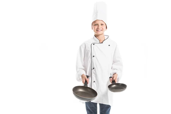 Smiling boy in white chef uniform and hat holding frying pans and looking at camera isolated on white — Stock Photo