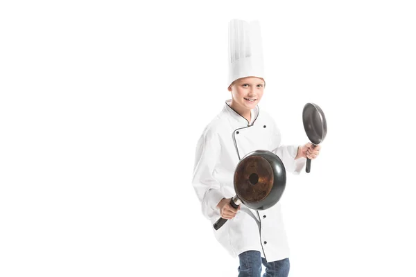 Smiling boy in white chef uniform and hat holding frying pans isolated on white — Stock Photo