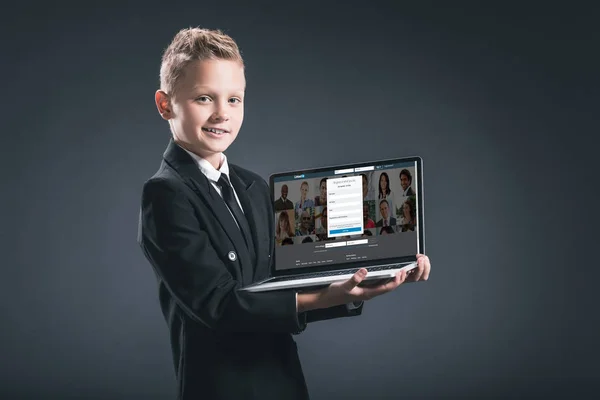 Smiling boy in businessman suit showing laptop with linkedin website on screen on grey background — Stock Photo