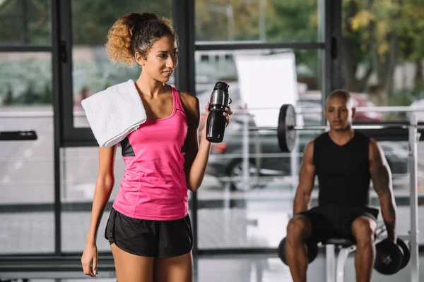 Bela afro-americana esportista segurando garrafa esporte enquanto seu namorado se exercitando atrás no ginásio — Fotografia de Stock