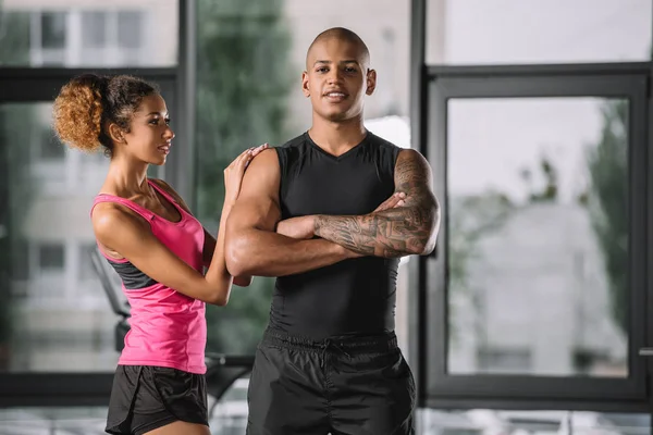 Muscular afro-americano masculino atleta posando com os braços cruzados, enquanto sua namorada de pé perto no ginásio — Fotografia de Stock