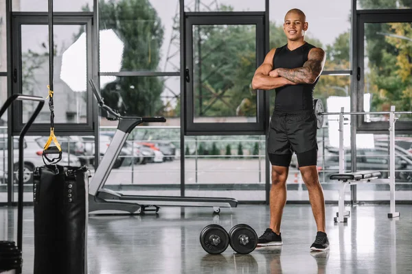 Athlète masculin tatoué posant avec les bras croisés près des haltères à la salle de gym — Photo de stock