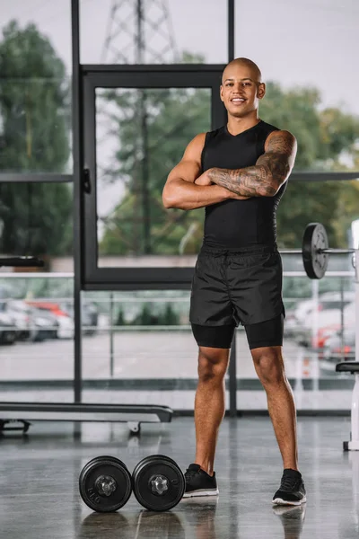 Apuesto deportista afroamericano posando con los brazos cruzados cerca de pesas en el gimnasio - foto de stock