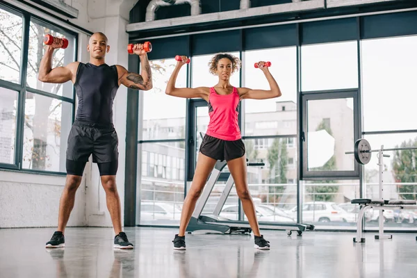 Sonriente joven afroamericano pareja de atletas haciendo ejercicio con mancuernas en el gimnasio - foto de stock