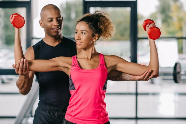 Sonriente afroamericano entrenador masculino ayudando a la deportista a hacer ejercicio con pesas en el gimnasio — Stock Photo