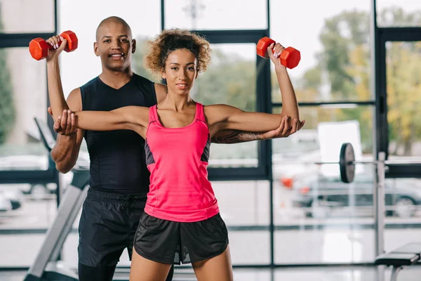 Joven afroamericano entrenador masculino ayudando a la deportista a hacer ejercicio con pesas en el gimnasio - foto de stock