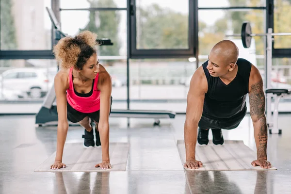 Sorrindo casal afro-americano de atletas olhando uns para os outros e fazendo prancha em tapetes de fitness no ginásio — Fotografia de Stock