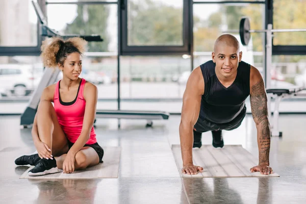 Guapo afroamericano atleta masculino haciendo tablón mientras su novia sentado cerca en el gimnasio — Stock Photo