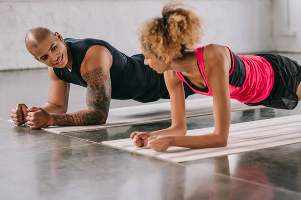 Jóvenes afroamericanos pareja de atletas mirándose y haciendo tablón en colchonetas de fitness en el gimnasio - foto de stock