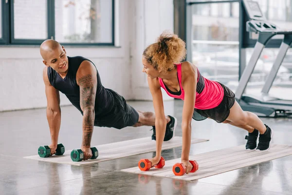 Couple of athletes looking at each other and exercising with dumbbells on fitness mats at gym — Stock Photo