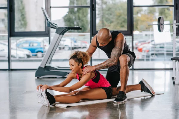 Tatuato africano americano maschio allenatore aiutare sportswoman a stretching in palestra — Foto stock