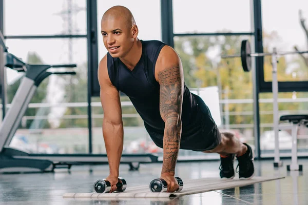 Bonito Africano americano desportista com tatuado mão fazendo flexões com halteres no tapete de fitness no ginásio — Fotografia de Stock