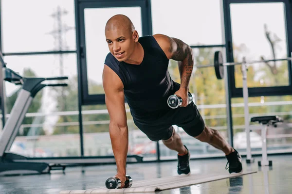 Sorridente sportivo afroamericano muscoloso che si allena con i manubri sul tappeto fitness in palestra — Foto stock