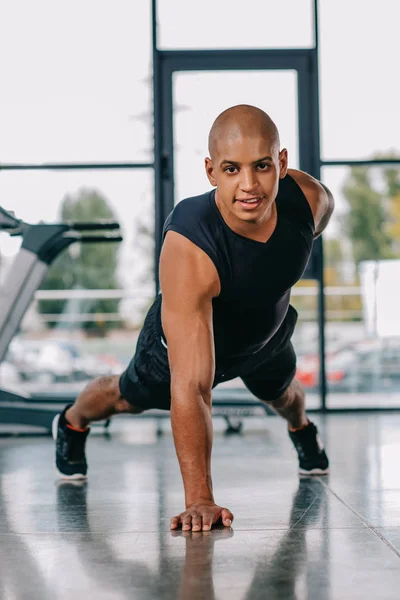 Deportista afroamericano mirando a la cámara y haciendo flexiones en una mano en el gimnasio - foto de stock