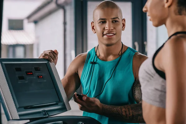 Selective focus of african american male trainer with timer looking at sportswoman running on treadmill at gym — Stock Photo