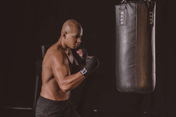 Giovane afroamericano maschio pugile in boxe bendaggio formazione con sacco da boxe in palestra — Foto stock
