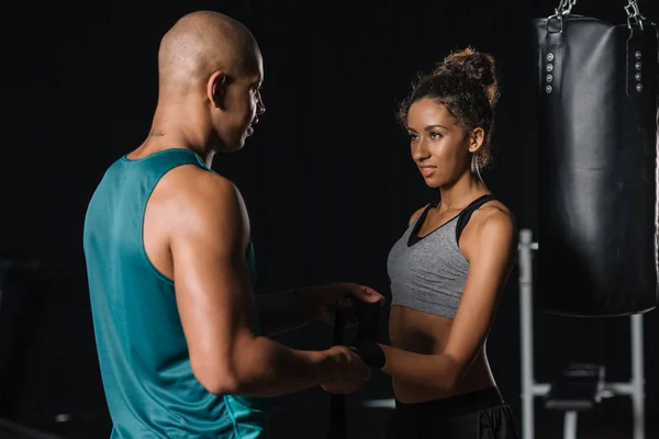 Smiling african american male trainer wrapping female boxer hand in boxing bandage at gym — Stock Photo