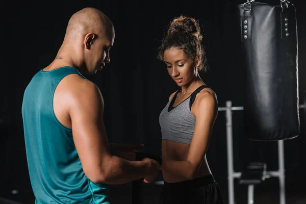 Seitenansicht eines afrikanisch-amerikanischen männlichen Trainers, der weibliche Boxerhände im Fitnessstudio in einen Boxverband wickelt — Stockfoto