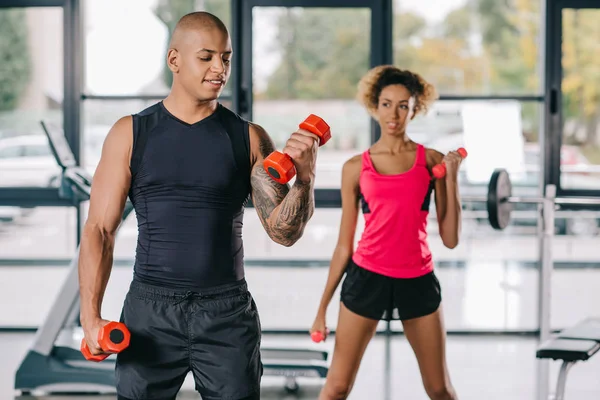 Sorridente afro-americano masculino treinador formação jovem mulher para o exercício com halteres no ginásio — Fotografia de Stock