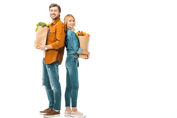 Alegre pareja con bolsas de compras llenas de productos de pie espalda con espalda aislado en blanco - foto de stock