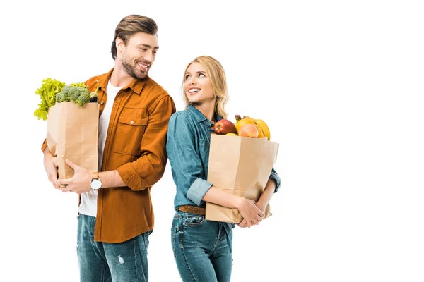 Pareja sonriente con bolsas de compras llenas de productos mirándose el uno al otro y parados espalda con espalda aislados en blanco - foto de stock