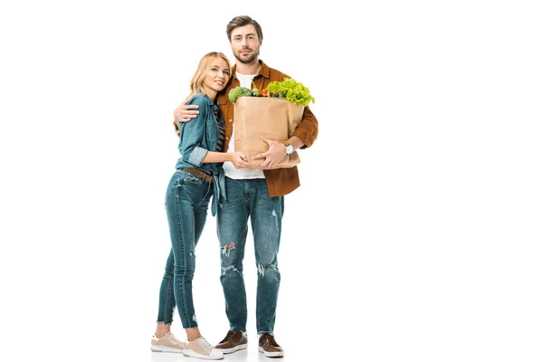 Alegre hombre sosteniendo bolsas de papel con productos y abrazando novia aislado en blanco - foto de stock