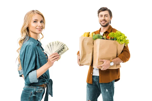 Молодая позитивная женщина показывает деньги в то время как ее парень стоит позади с продуктами в мешках для покупок изолированы на белом — стоковое фото