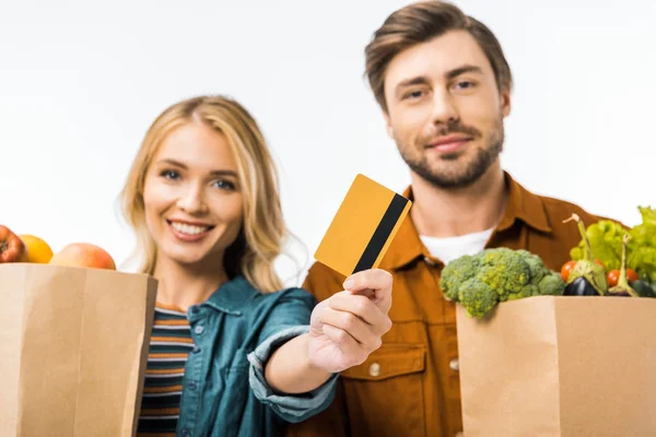 Foco seletivo da mulher mostrando cartão de crédito, enquanto seu namorado de pé perto com sacos de compras cheios de produtos isolados no branco — Fotografia de Stock