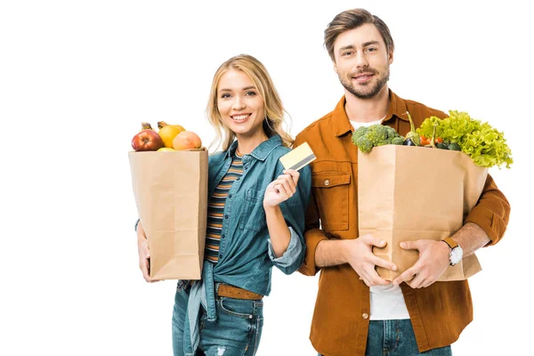 Sonriente hermosa chica mostrando tarjeta de crédito mientras su novio de pie cerca con bolsas de compras llenas de productos aislados en blanco - foto de stock