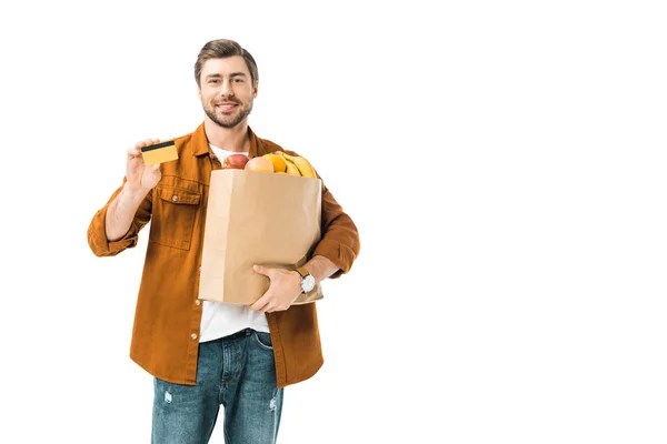 Hombre positivo con bolsa llena de productos que muestran tarjeta de crédito aislada en blanco - foto de stock