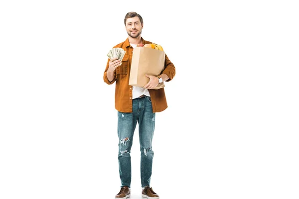 Человек, показывающий деньги и держащий бумажный пакет, полный продуктов, изолированных на белом — стоковое фото