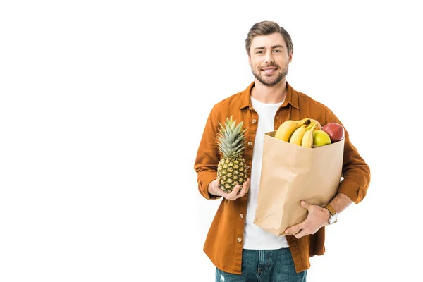 Hombre sonriente sosteniendo piña y bolsa de papel llena de productos aislados en blanco - foto de stock