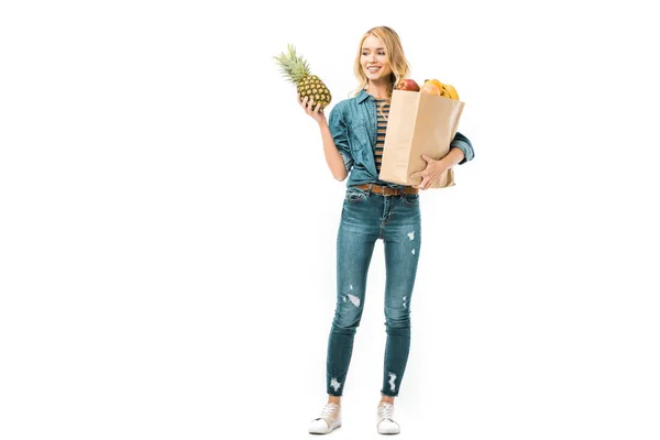 Smiling young woman looking at pineapple and holding paper bag with products isolated on white — Stock Photo