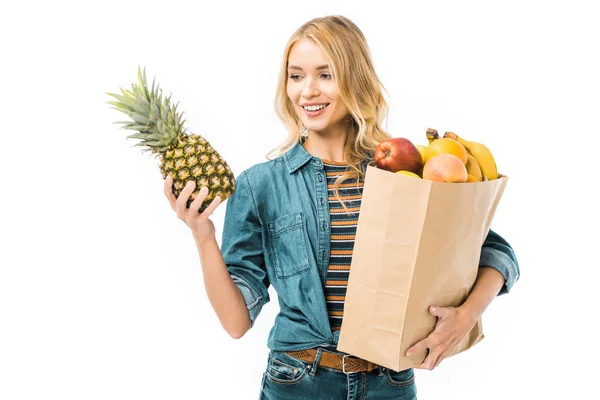 Mujer alegre mirando piña y sosteniendo bolsa de papel con productos aislados en blanco - foto de stock
