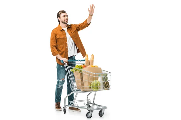 Uomo sorridente che ondeggia a mano e in piedi vicino al carrello della spesa con prodotti in sacchetti di carta isolati su bianco — Foto stock