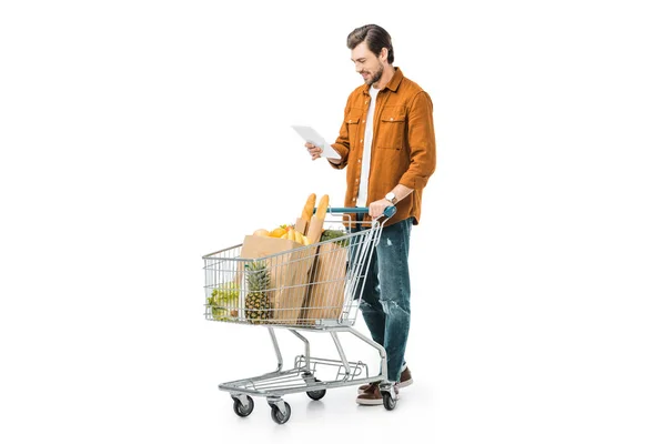 Handsome man checking shopping list on digital tablet and carrying shopping trolley isolated on white — Stock Photo