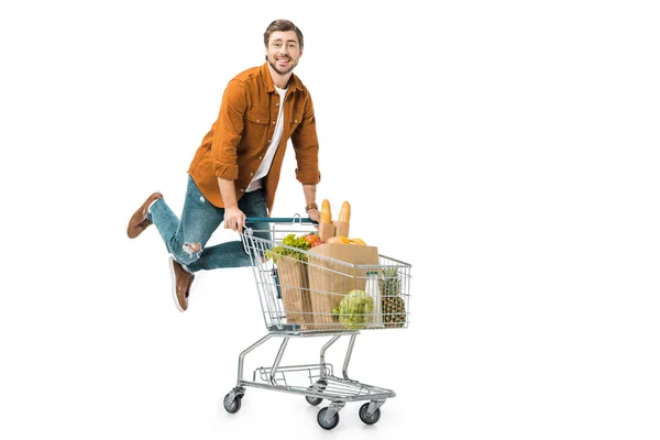 Hombre positivo saltando y llevando carrito de compras con productos en bolsas de papel aisladas en blanco - foto de stock