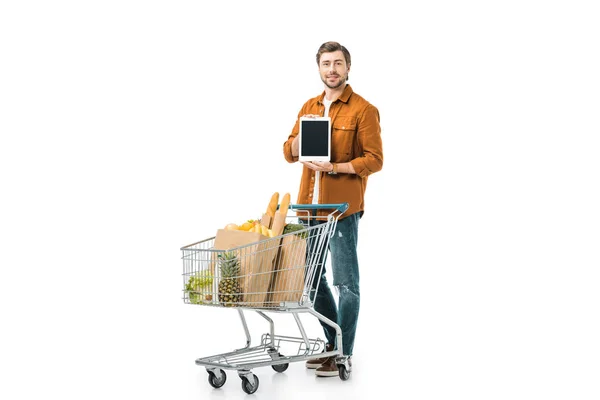 Hombre feliz mostrando tableta digital con pantalla en blanco cerca del carro de la compra con bolsas de papel aisladas en blanco - foto de stock