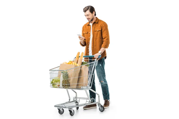 Felice uomo bello che trasporta carrello della spesa con prodotti in sacchetti di carta e utilizzando smartphone isolato su bianco — Foto stock